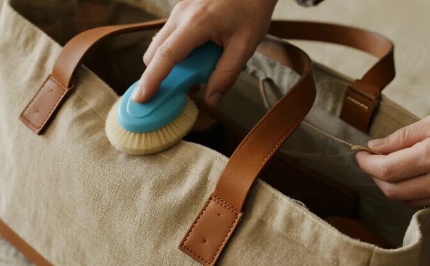 A woman holds a brush in one hand and a bag in the other, standing with a focused expression.