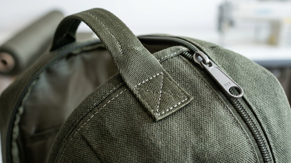Macro photograph of even double stitching, reinforced handle attachment, and a quality metal zipper pull on textured green RPET fabric